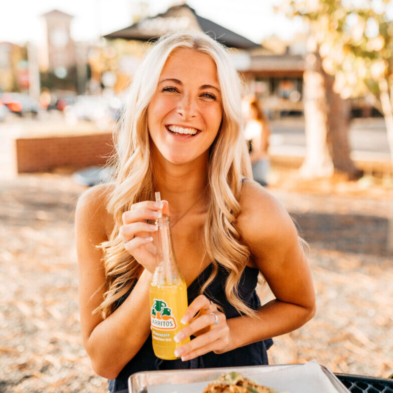 A smiling woman with long blonde hair sits outdoors, holding a bottle of Jarritos pineapple soda with a straw. She is wearing a sleeveless top and enjoying a meal on a sunny day.