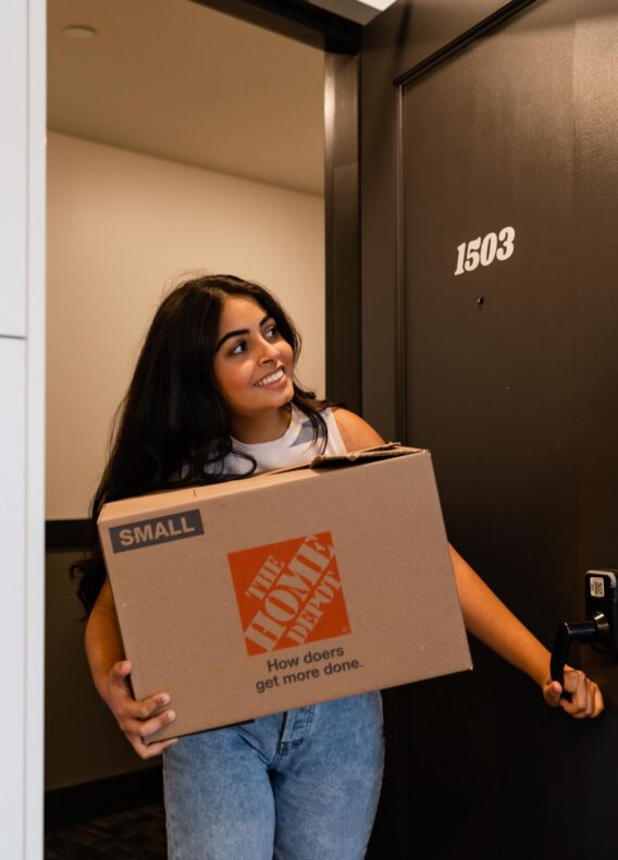A woman smiles while holding a cardboard moving box labeled “The Home Depot” and stands in the doorway of an apartment near Georgia Tech with the number 1503.