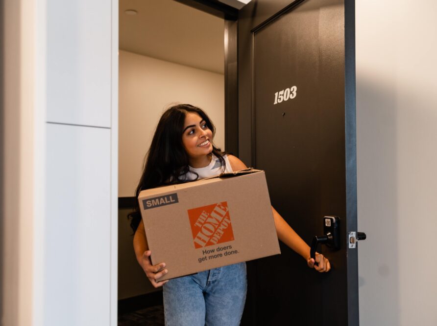 A woman smiles while holding a cardboard moving box labeled “The Home Depot” and stands in the doorway of an apartment near Georgia Tech with the number 1503.